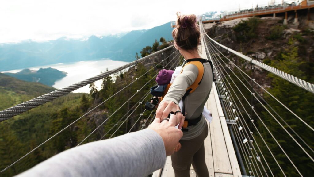 A parent using a supportive baby carrier while walking across a suspension bridge in a mountainous landscape, demonstrating the freedom of babywearing in the outdoors.
